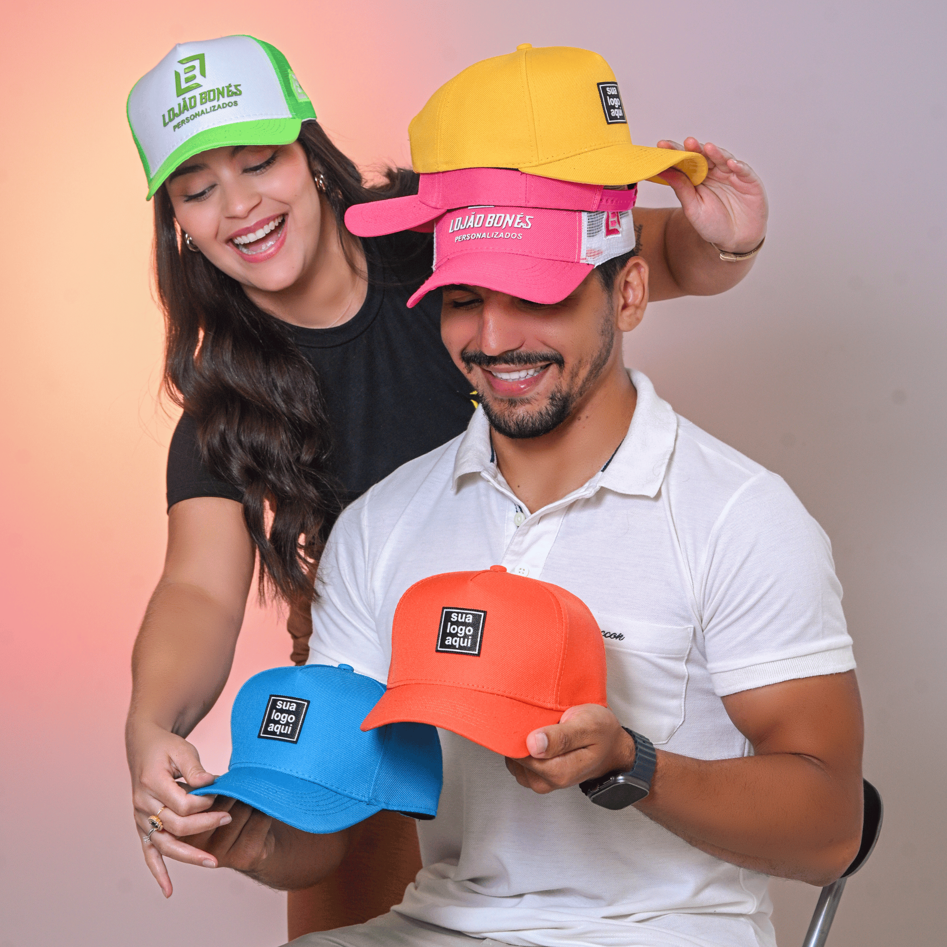 Casal sorrindo segurando bonés personalizados em diversas cores.
