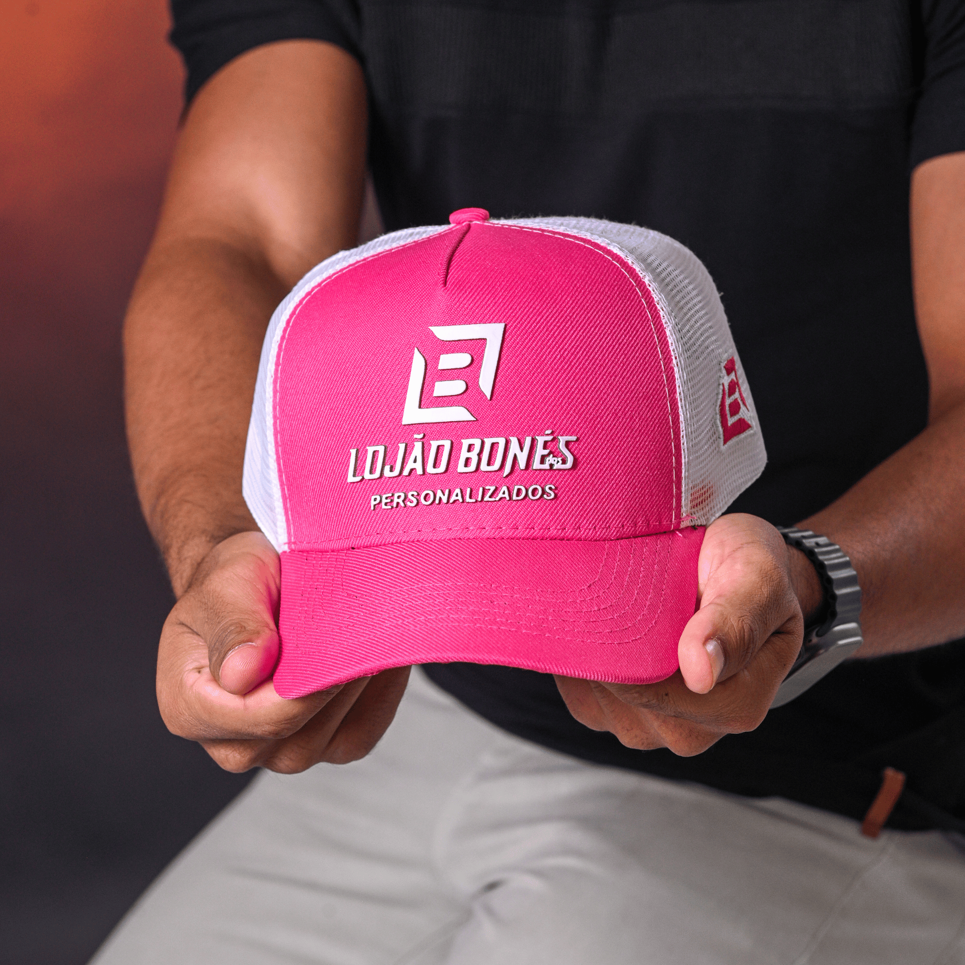 Mão segurando boné trucker rosa com tela branca e logo em detalhe.