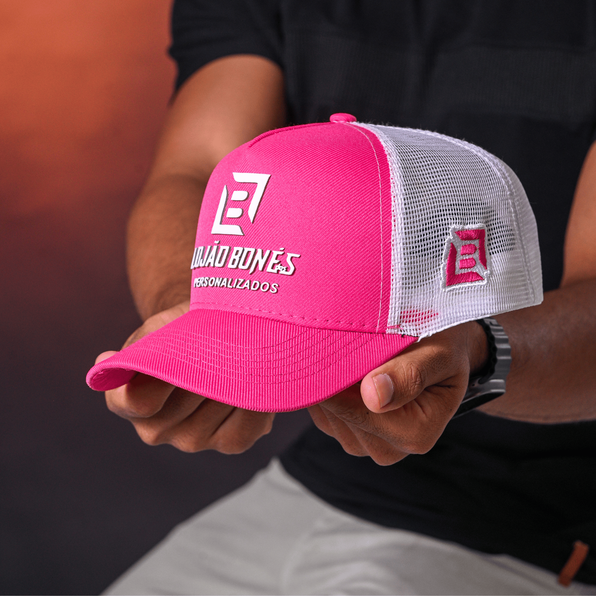 Modelo segurando um boné trucker personalizado com aba curva rosa e frente branca com o logo do Lojão dos Bonés.
