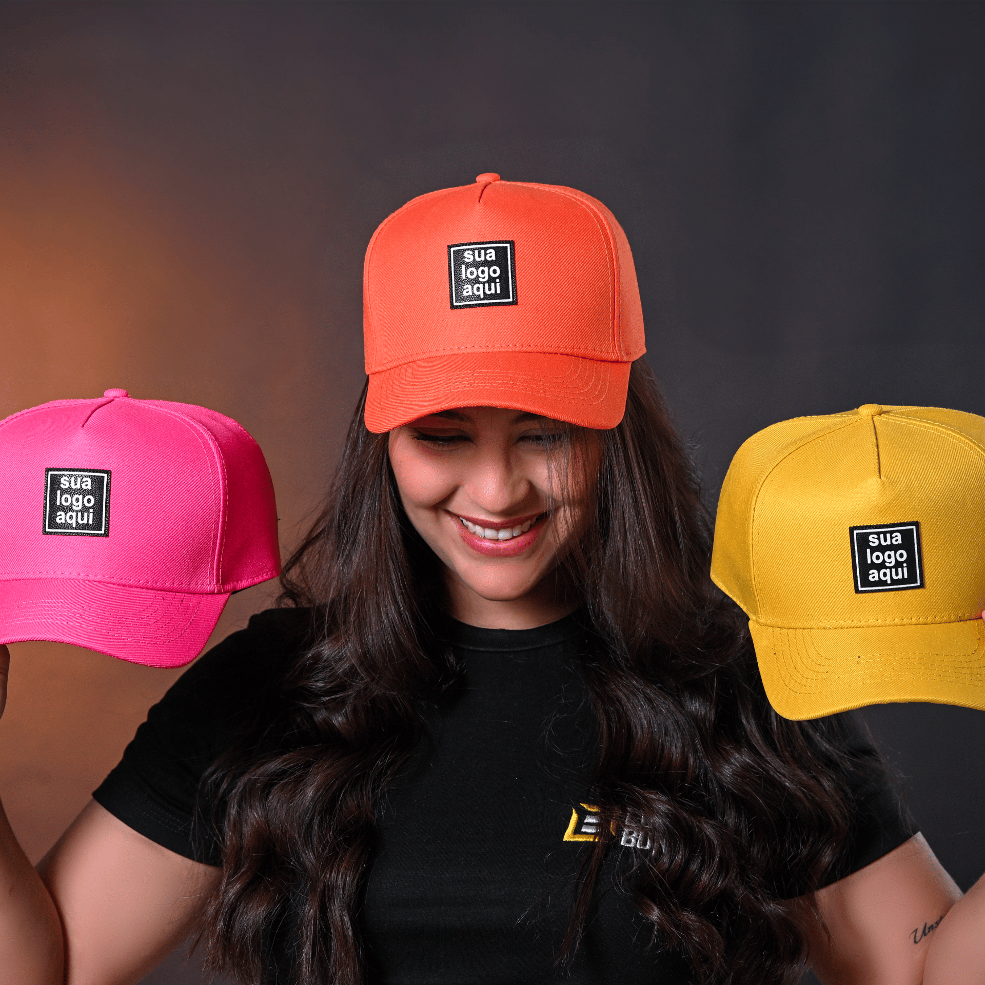 Modelo feminina sorrindo usando boné laranja, com bonés rosa e amarelo ao fundo.